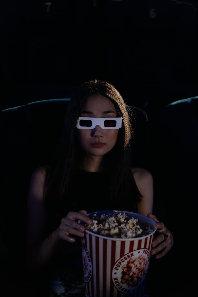 An adult woman sitting in a dark movie theater wearing 3D glasses and holding popcorn.